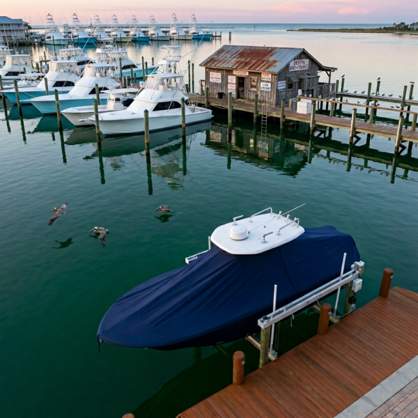 Covered speedboat on a lift at a wooden dock in a calm harbor, with white boats and a weathered shack in the background.