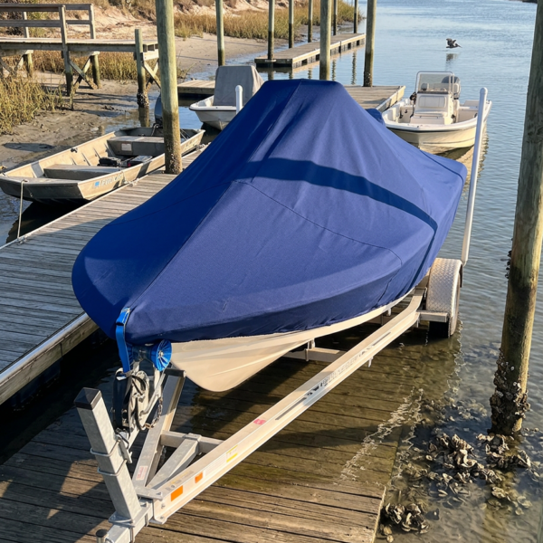 Blue-covered boat on a trailer docked at a wooden pier, with calm canal water, pilings, and other boats in the background.