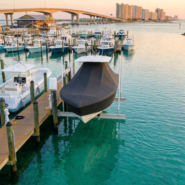 Boats docked at a wooden marina with a covered boat on a lift and turquoise water, city skyline and bridge in the distance.]