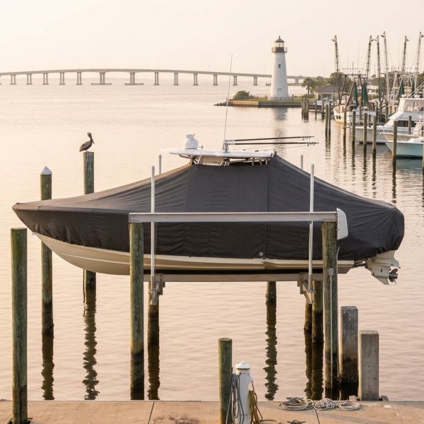 A boat covered with a dark tarp sits on a lift at a quiet marina with a lighthouse in the distance.