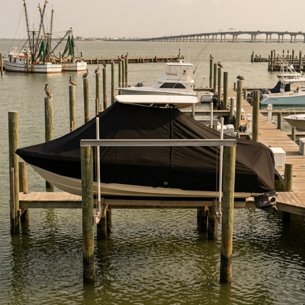 Covered motorboat on a wooden dock at a marina, with other boats and a distant bridge in the background.