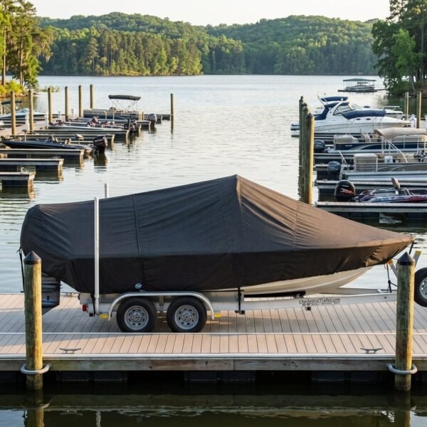 Covered boat on a dual-axle trailer parked on a wooden dock at a calm marina, with other boats nearby and trees in the distance.