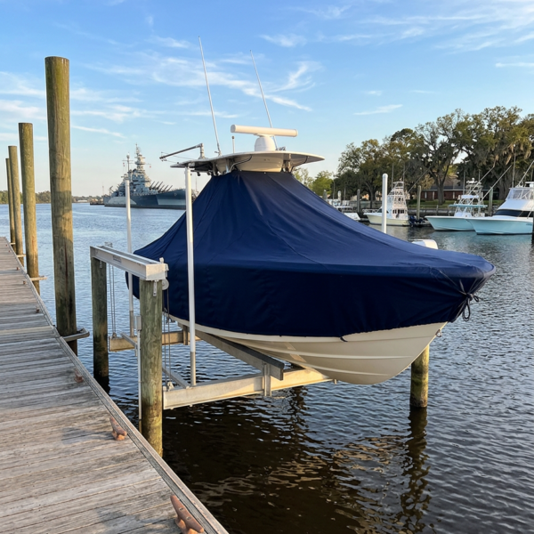 Boat covered with a dark blue tarp on a boat lift at a wooden marina dock, calm water and blue sky nearby.