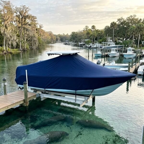 A blue-covered boat docked at a wooden marina with calm water and visible manatees resting beneath the surface nearby.