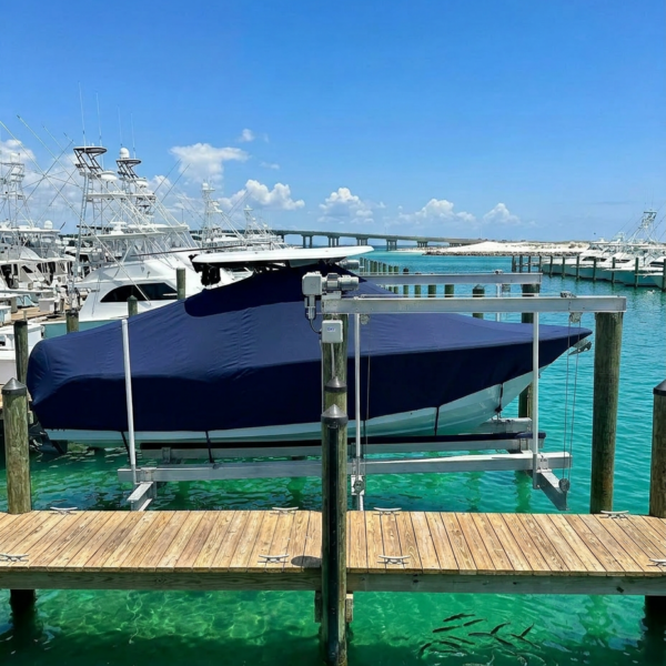 Boats moored at a marina; a large boat with a dark blue cover sits on a lift at a wooden dock above turquoise water under a bright blue sky.