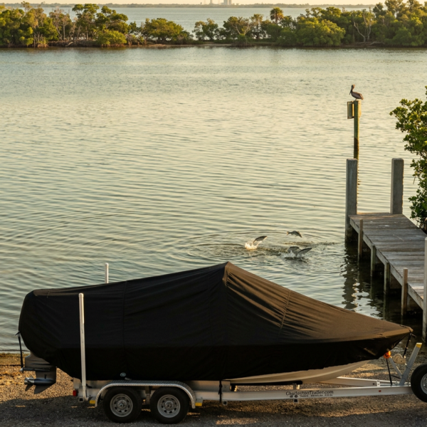 Boat covered with a black cover on a trailer by a rocky shore, next to a wooden dock on calm water.