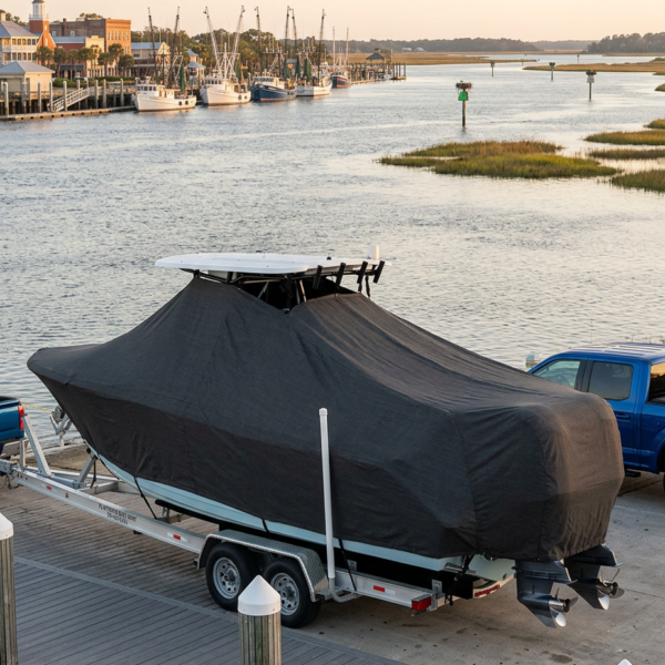 Boat on a trailer at a marina, covered with a black protective cover, with water and moored boats in the background.