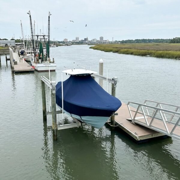 Boat dock with a blue-covered motorboat on a lift, calm water and marshlands in the distance; city skyline on the horizon.