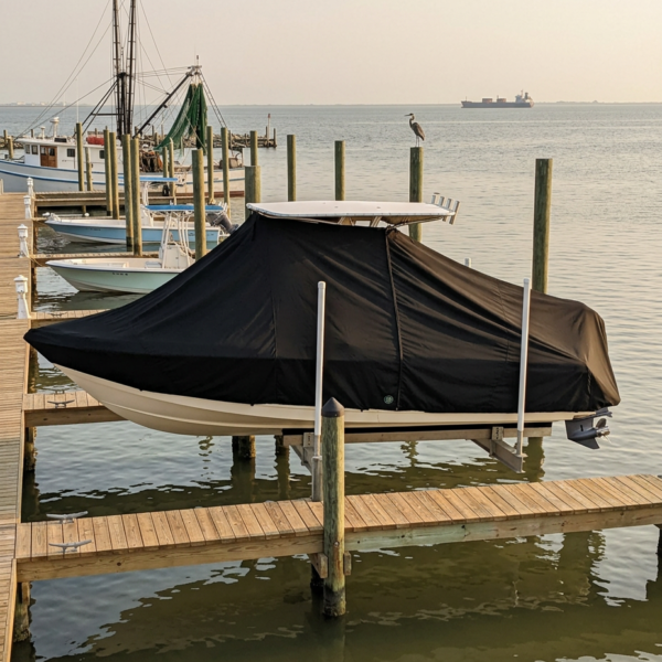Boat covered with a black tarp on a wooden dock by calm water, seagull perched on a piling in the background