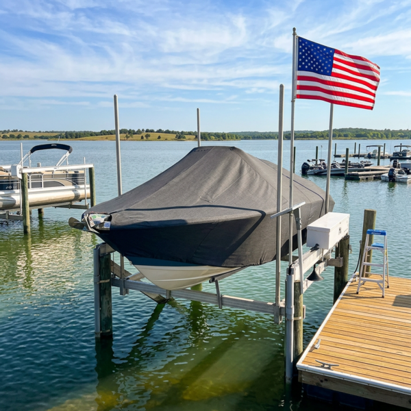 Boat covered with a dark boat cover on a lift at a wooden dock, American flag waving nearby over calm water under a blue sky.
