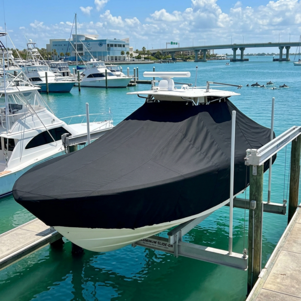 White motorboat docked in a marina with a black cover over the hull, moored to a floating dock amid blue-green water and other boats. In the background, a bridge and coastal buildings under a sunny sky with scattered clouds.