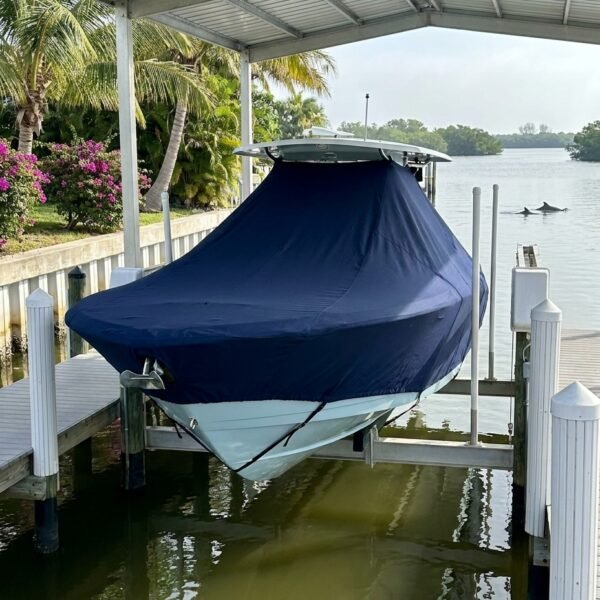 Blue-covered boat in a covered dock slip by calm water with palm trees and flowers nearby, marina setting.