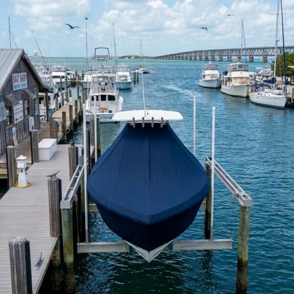 Blue-covered boat on a lift at a marina with wooden docks and several white yachts in the background.