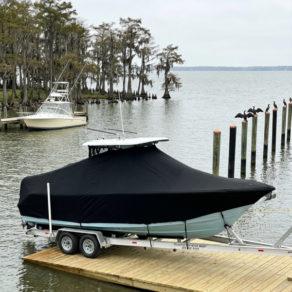 Boat covered with a black tarp on a dual-axle trailer at a wooden dock by calm water.