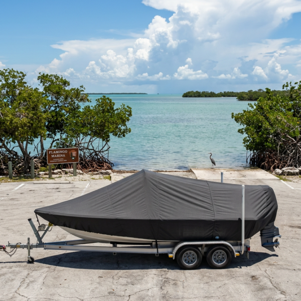 Covered boat on a dual-axle trailer parked on cracked pavement beside a marina, with turquoise water and mangrove trees in the background.