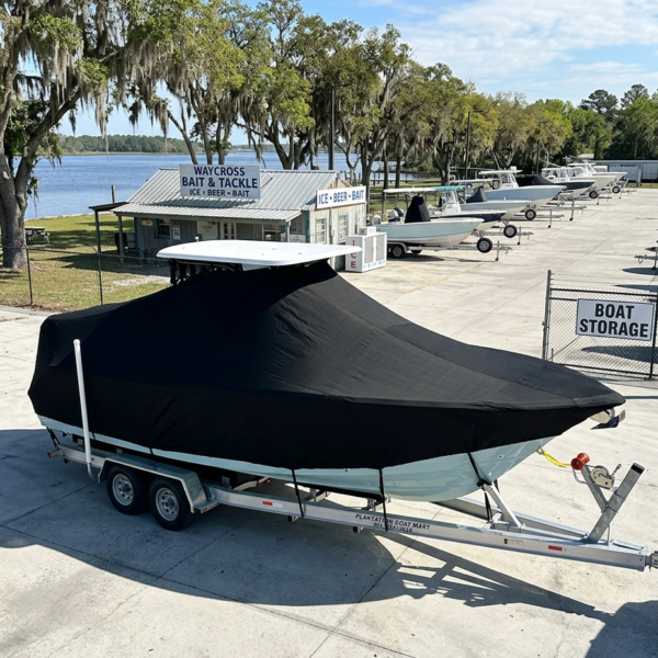 Covered boat on a trailer in a boat storage lot by the water, with trees and a marina shop in the background.