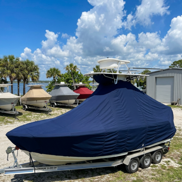 Blue-covered boat on a triple-axle trailer in a boat yard by the water and palm trees behind it.