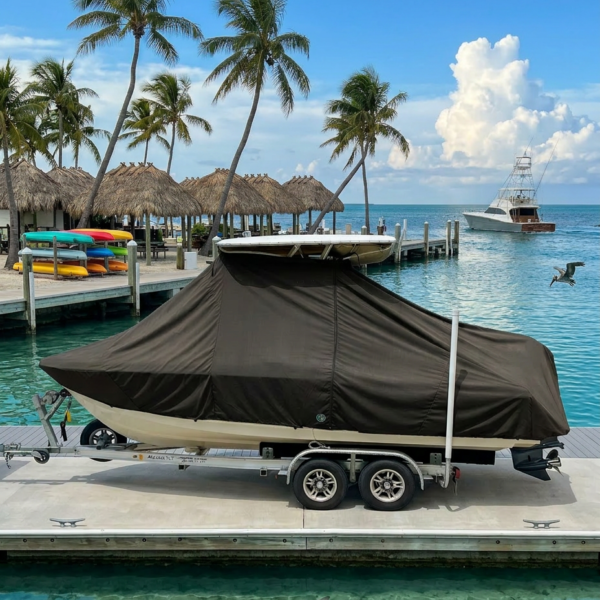 Boat on a trailer covered with a brown tarp at a sunlit marina dock, turquoise water and palm trees in the background.