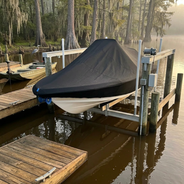 Boat covered with a black tarp on a boat lift at a wooden dock by a calm, muddy lake with trees in the background.