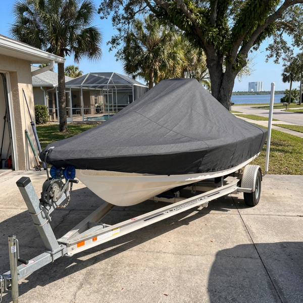 Covered boat on a dual-axle trailer parked in a suburban driveway with palm trees and a lake in the background