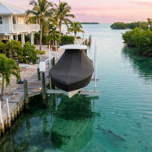 A dockside scene with a covered boat lifted on a lift over clear turquoise water, palm trees, and a pink sunset sky.