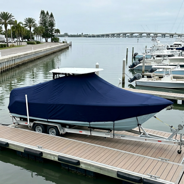 Blue-covered boat on a dual-axle trailer docked at a marina with calm water and palm trees in the background.