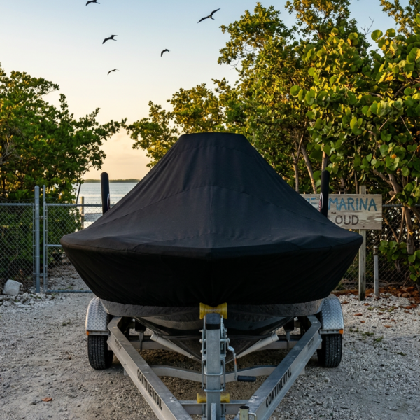 Covered boat on a trailer in a gravel lot with a chain-link fence and trees at sunset/sunrise background