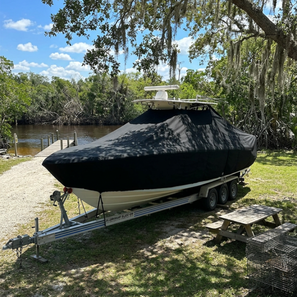 Powerboat on a triple-axle trailer covered with a black tarp, parked near a river under trees and sunshine.