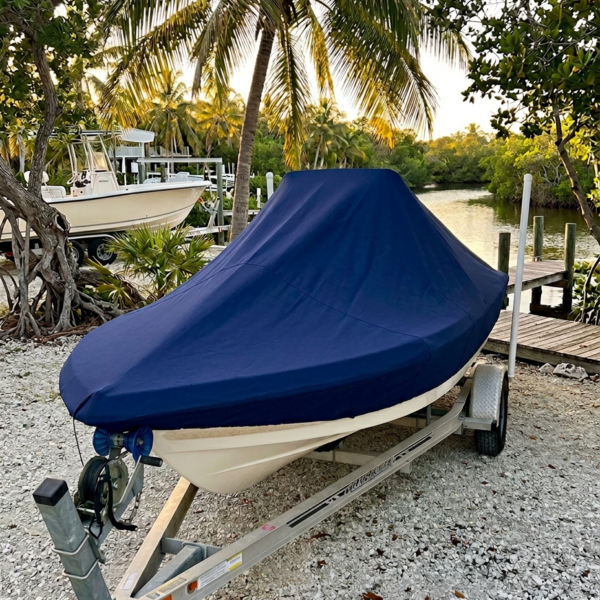 Boat on a trailer with a blue cover parked on a gravel area beside a canal and palm trees under a tropical sky.