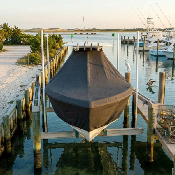 A covered boat on a lift at a marina, with calm water, pilings, a white heron on a post, and moored boats in the background.