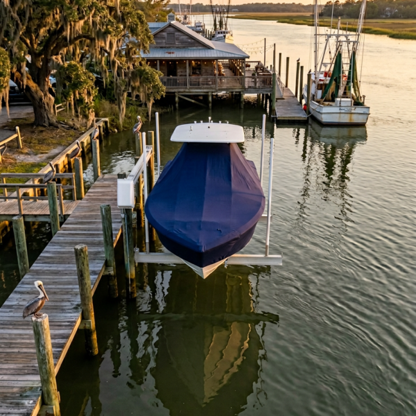 Blue-covered boat on a boat lift at a wooden dock, with a pelican on a piling and calm water reflecting the scene at sunset.