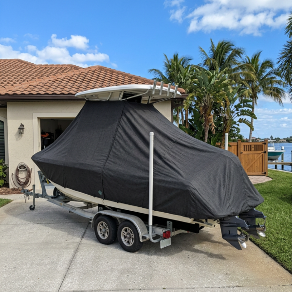 Boat on a dual-axle trailer in a driveway, covered by a black boat cover, with palm trees and a tiled-roof house in the background.