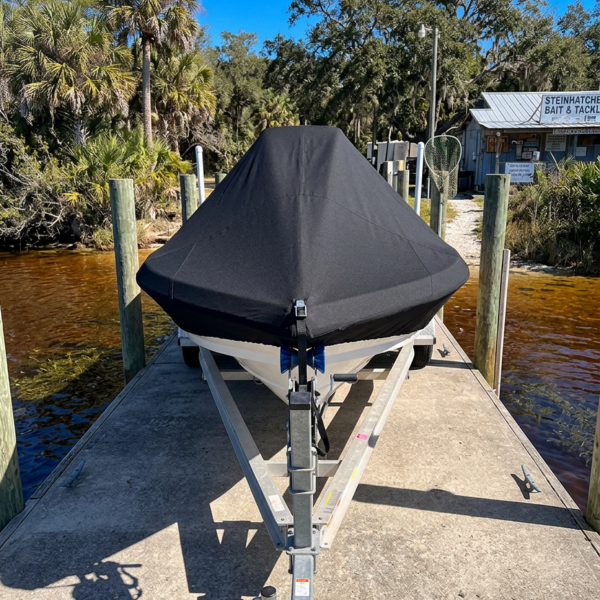 A boat on a trailer with a black cover parked on a dock beside brown water and tropical trees under a blue sky.