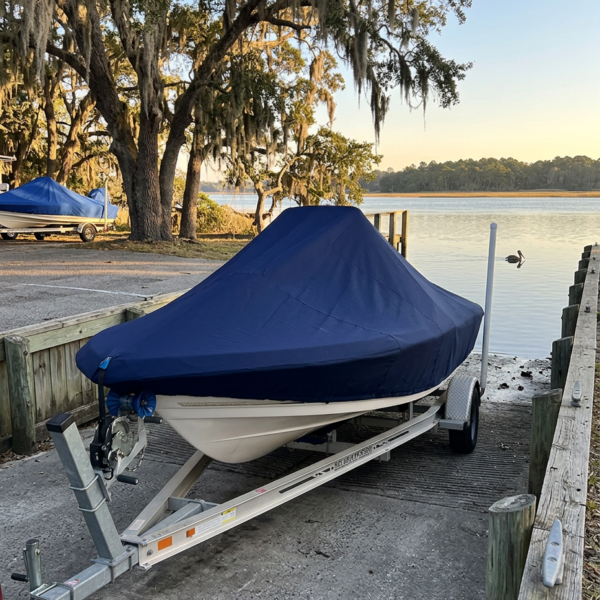 Boat on a trailer covered with a dark blue boat cover by a lakeside dock, with trees and calm water in the background