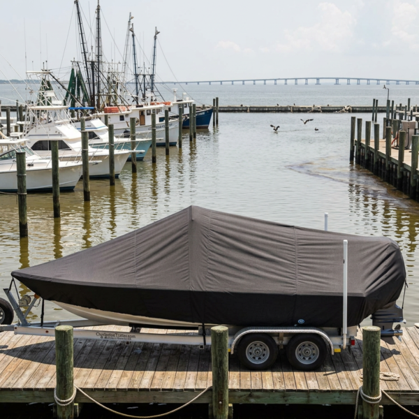 Boat covered with a gray tarp on a dual-axle trailer at a wooden marina dock, with moored boats to the left and a distant bridge over the water.