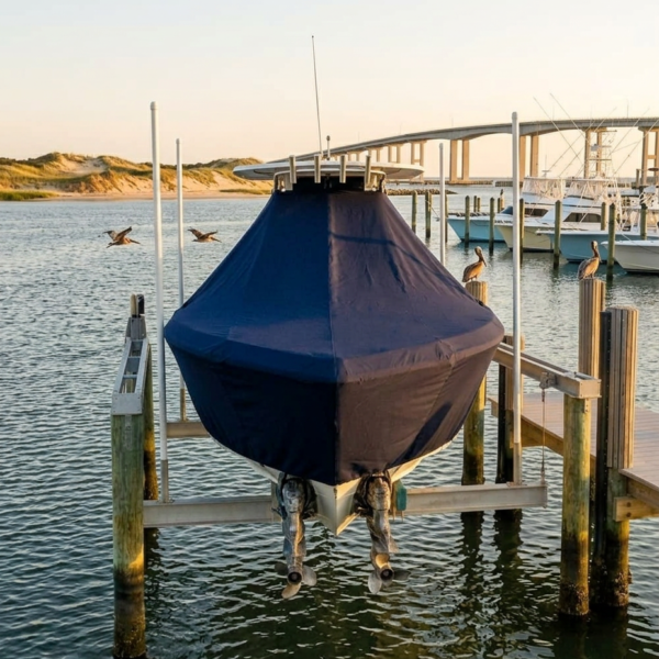 Blue-covered boat on a lift at a marina with pilings and calm water, coastal dunes in the background.