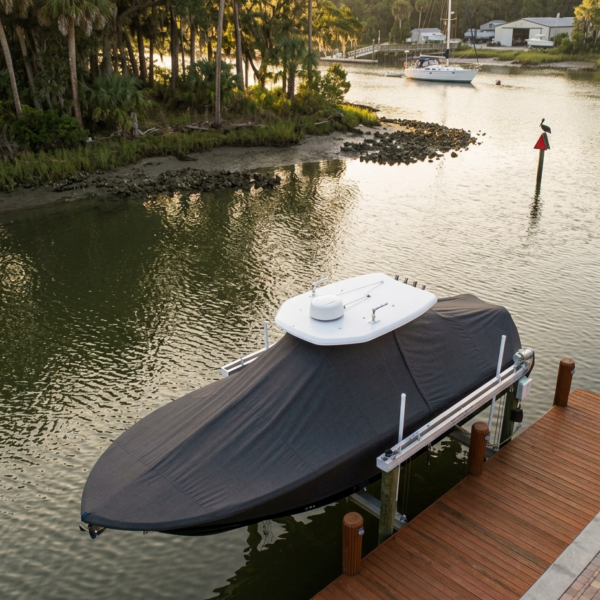 Covered speedboat moored at a wooden dock along a calm inlet with palm trees and a distant sailboat in the background
