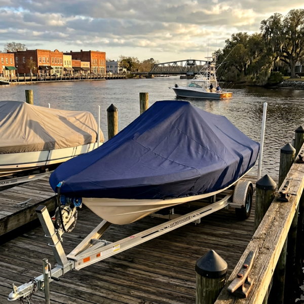 Boat on a trailer covered with a dark blue tarp, docked at a wooden marina with calm water and a distant bridge.