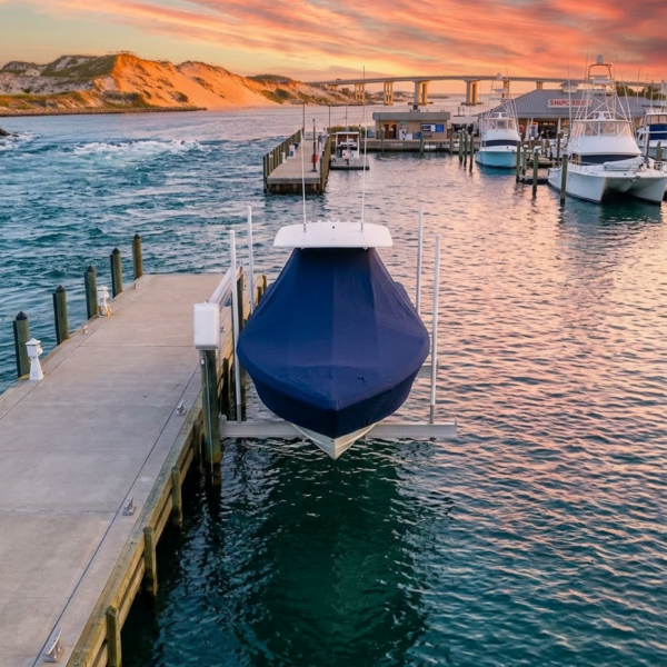 Blue-covered boat on a boat lift at a marina at sunset, with calm turquoise water and other boats in the background.