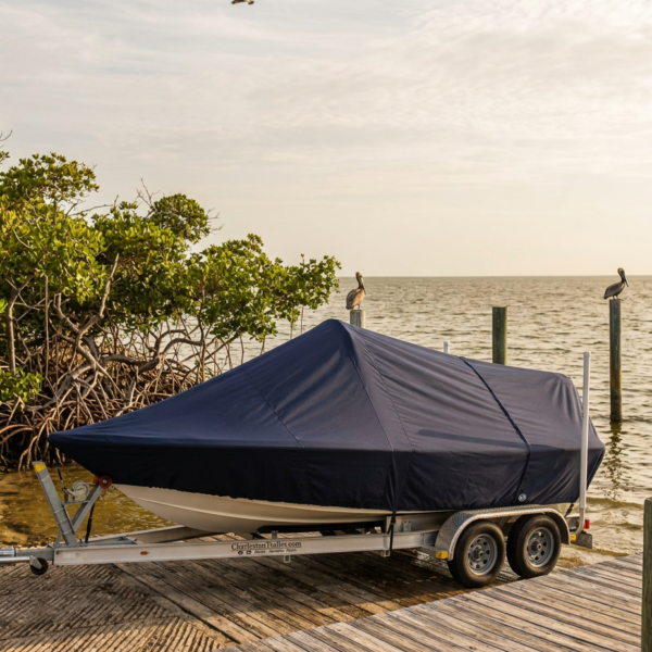 Boat on a dual-axle trailer covered with a dark tarp, parked on a wooden dock by the water.